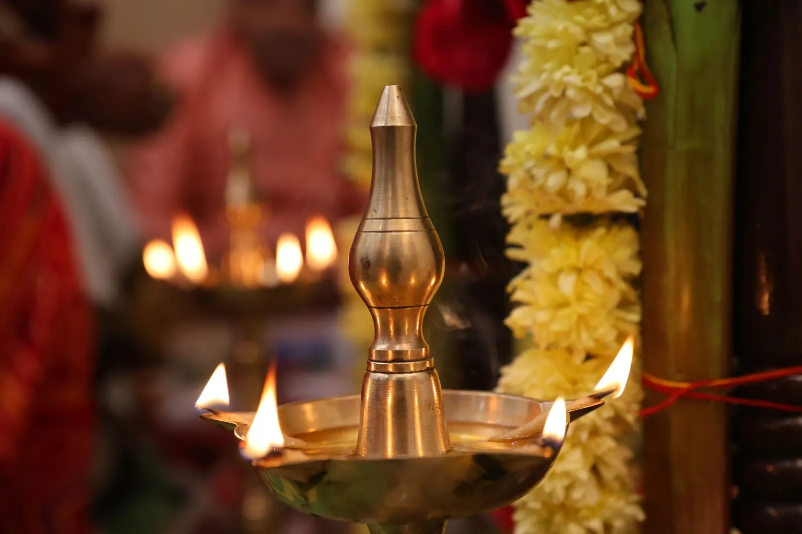 A lit brass oil lamp with several flames beside a garland of pale yellow flowers