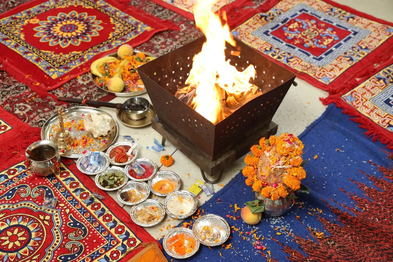 A havan fire in a metal kund, with puja thalis of offerings and a marigold-decked kalash on embroidered mats