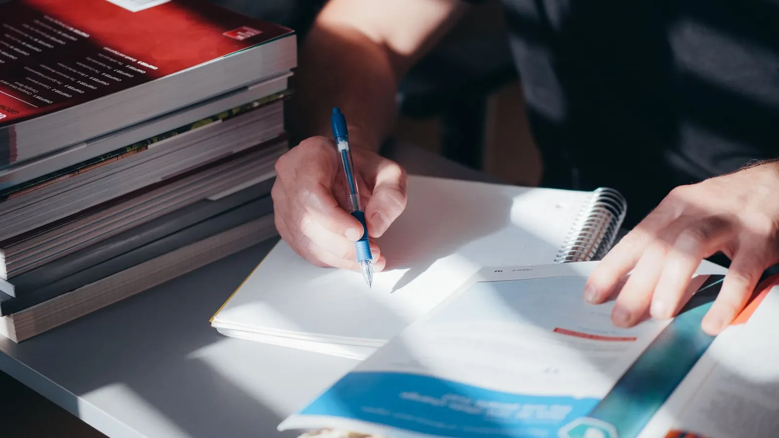 Hands writing in a spiral notebook beside a stack of textbooks on a sunlit desk