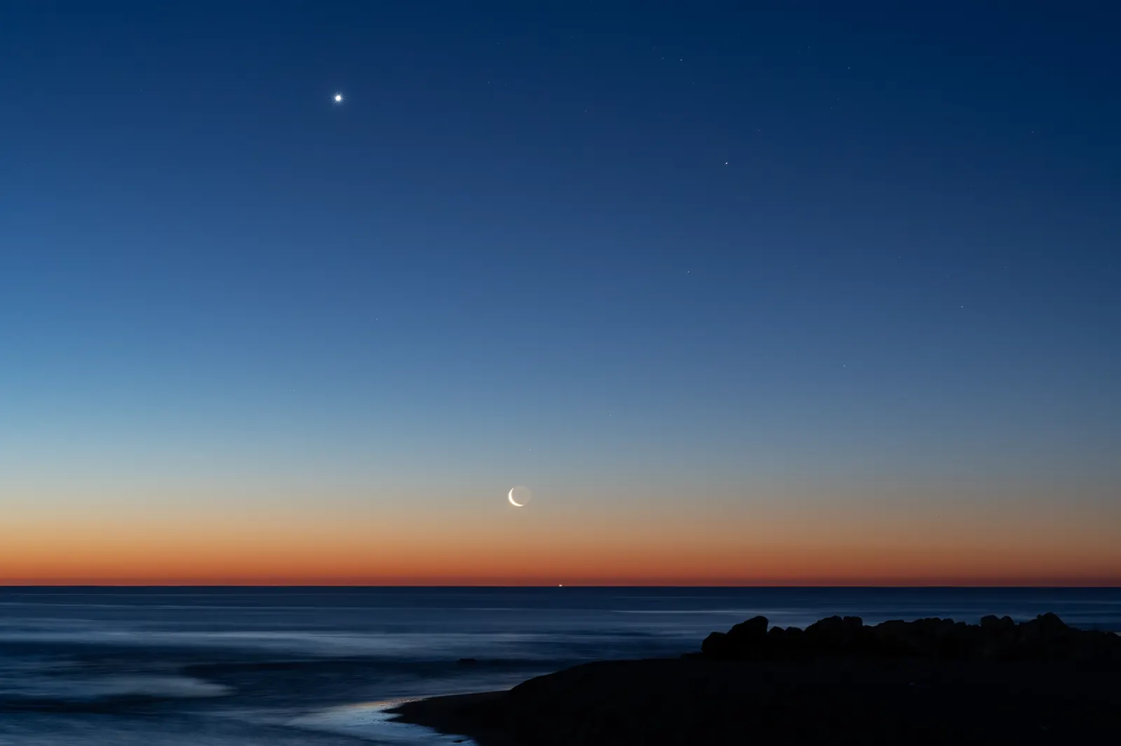 A thin crescent moon and two bright planets in a twilight sky over the sea