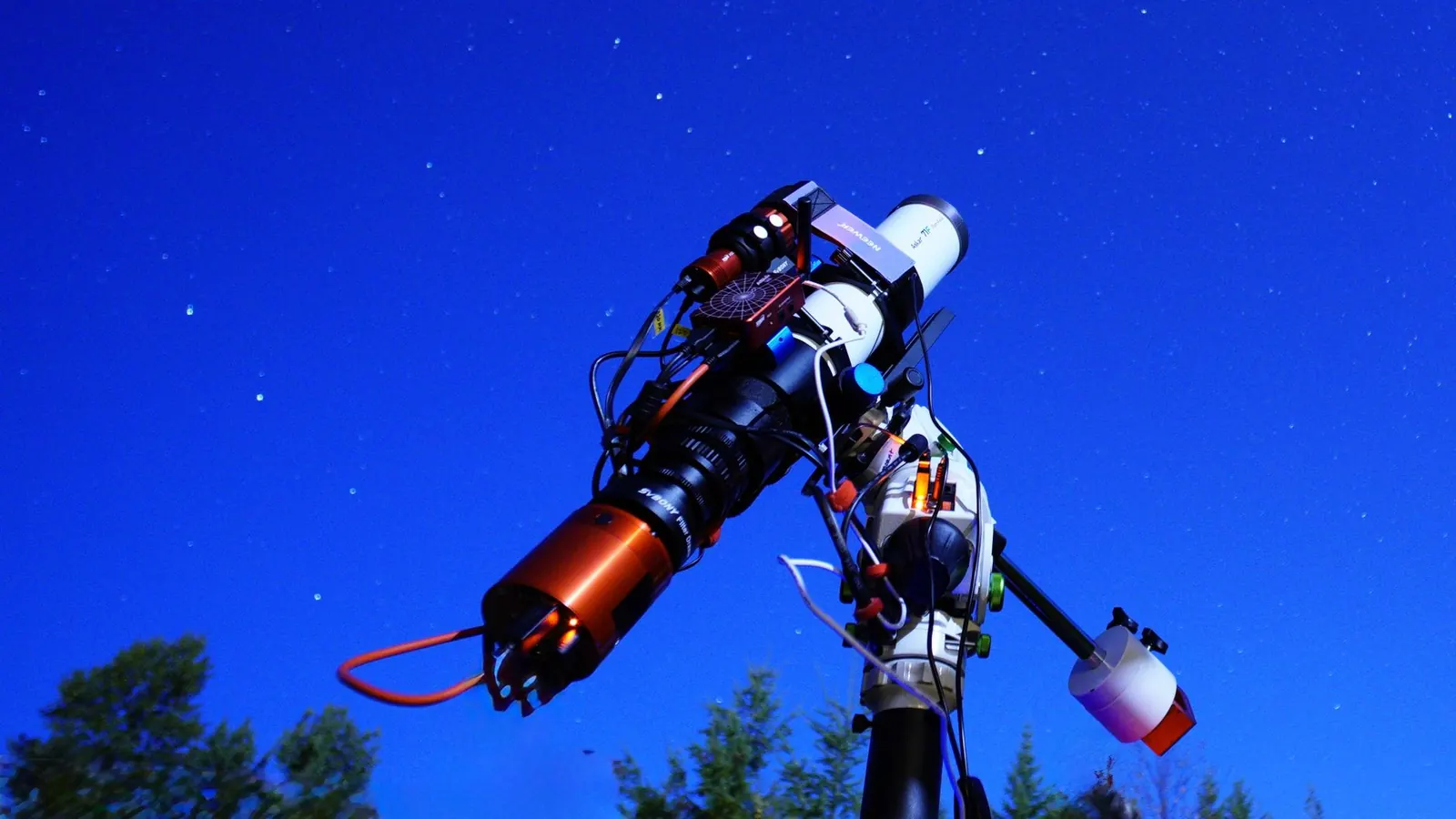 A telescope with a camera on a mount, pointed at a starry night sky above treetops