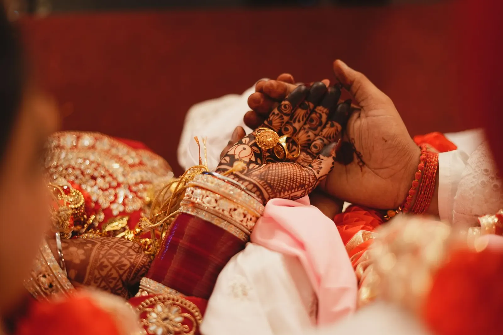A groom's hand holding a bride's hennaed hand, with red bangles and gold jewellery, at an Indian wedding