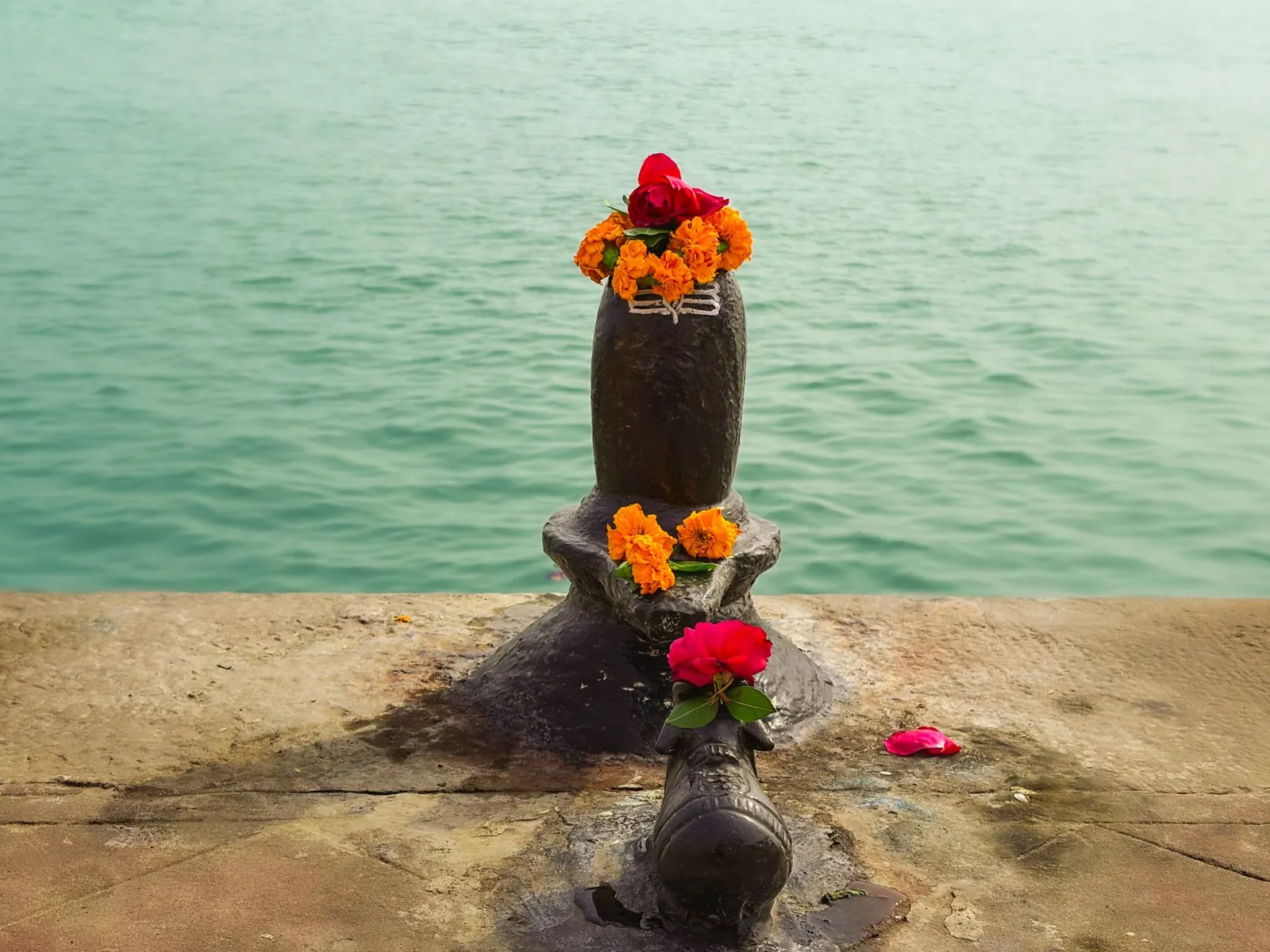 A stone Shiva lingam topped with marigolds and a red flower on a riverside ghat, with a small Nandi in front