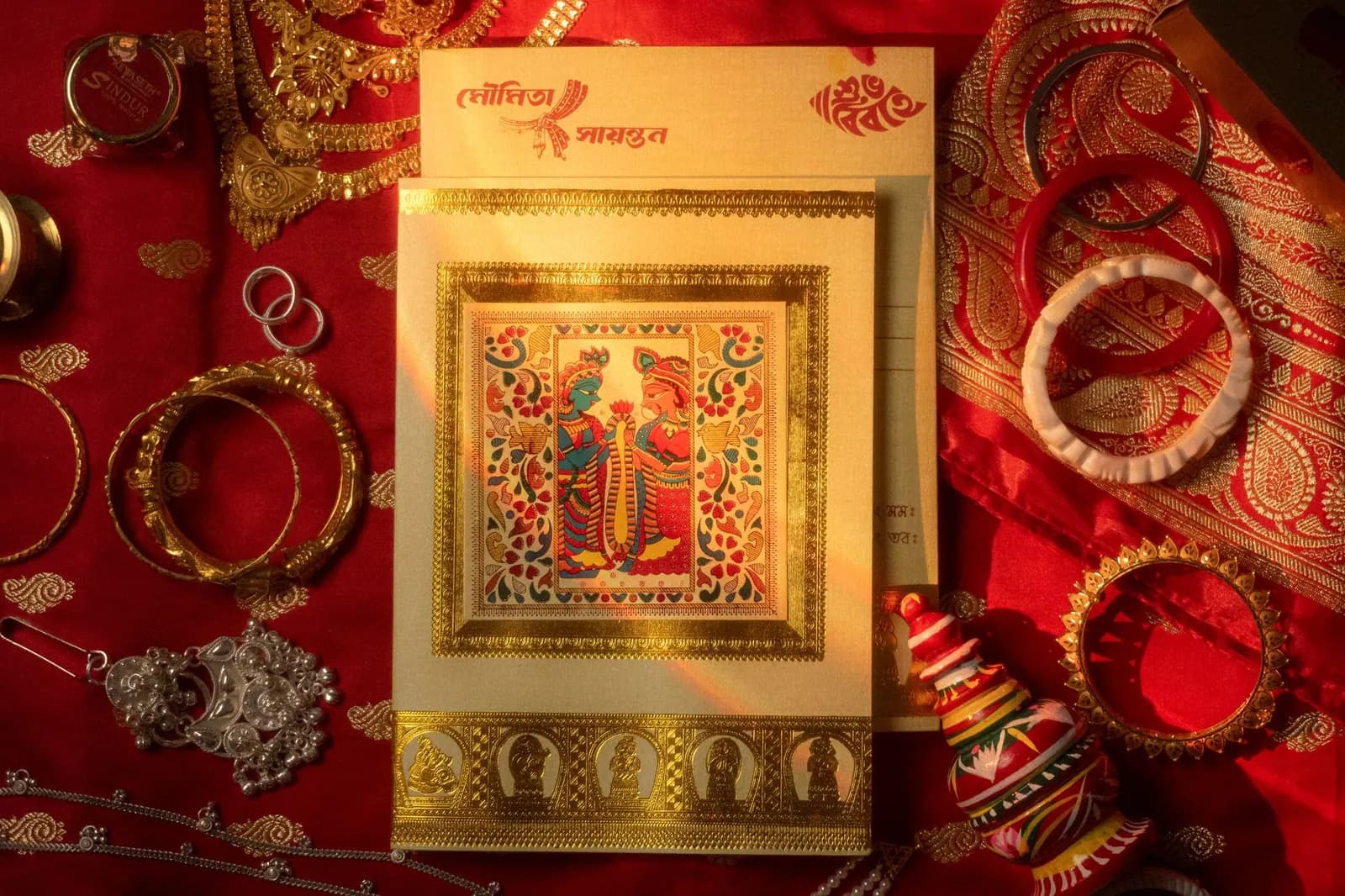 A Bengali wedding invitation card on red cloth, surrounded by bangles and gold jewellery