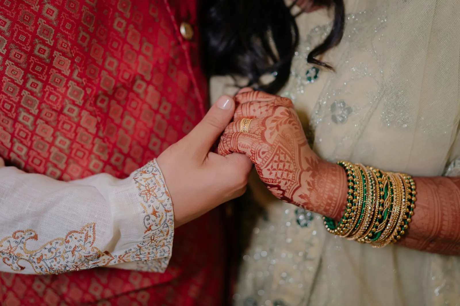 A couple holding hands in Indian wedding clothes, the bride's hand covered in mehndi and bangles