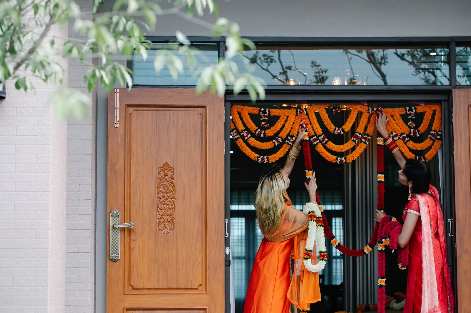 Two women hang a marigold garland across a home's front entrance beside a carved wooden door