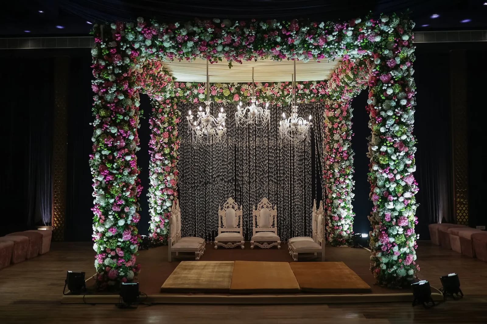 An empty wedding mandap framed with pink and white flowers, with chandeliers and four chairs
