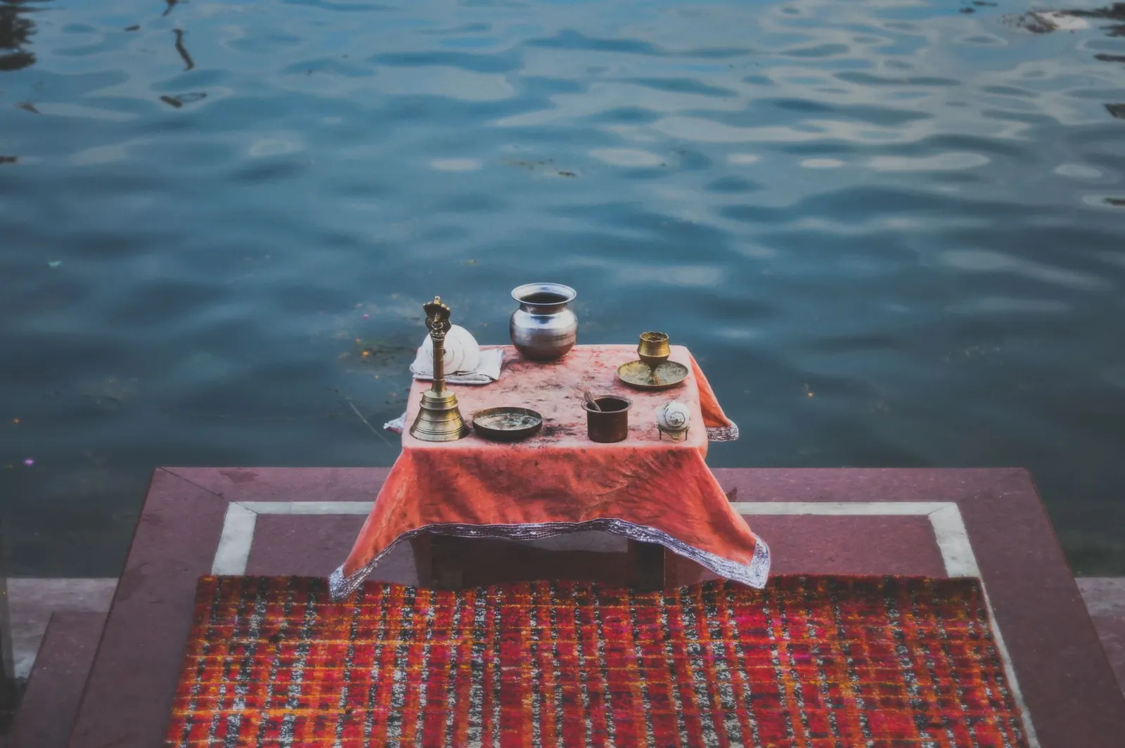Brass lota, bell and puja vessels on a cloth-covered stool at a riverside ghat, set for a ritual