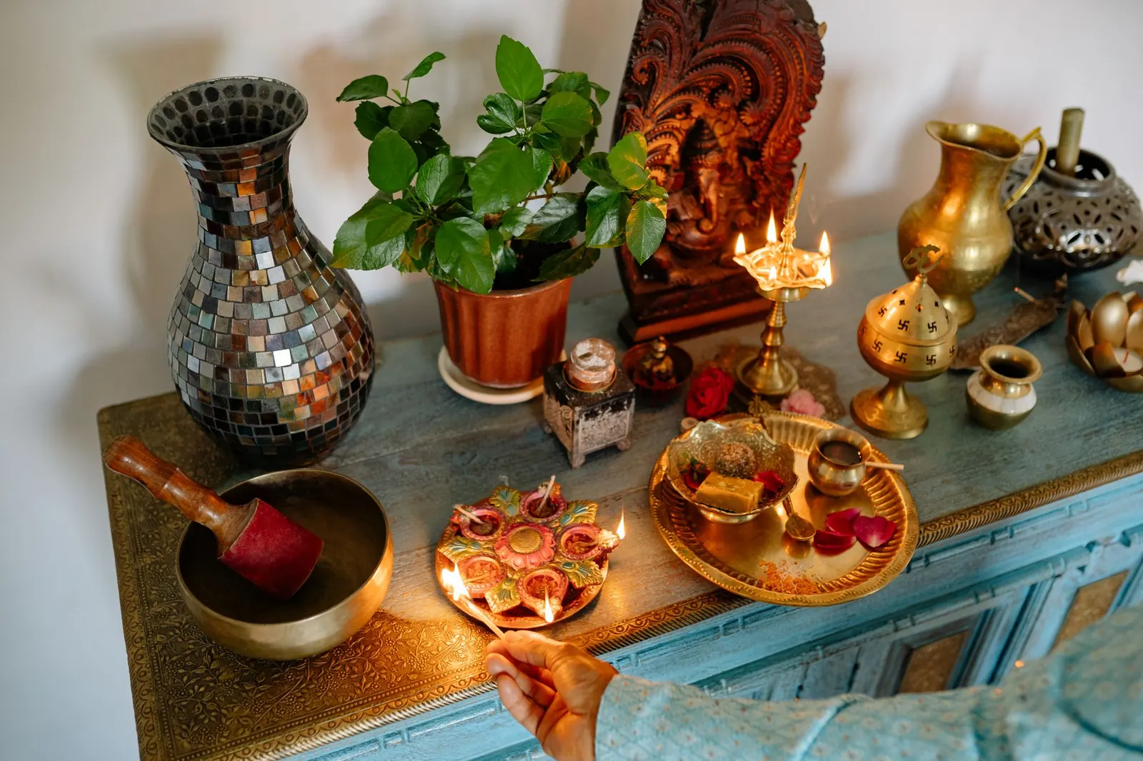 A home puja shelf against a wall with a carved Ganesha panel, a lit brass lamp, small diyas and a brass plate