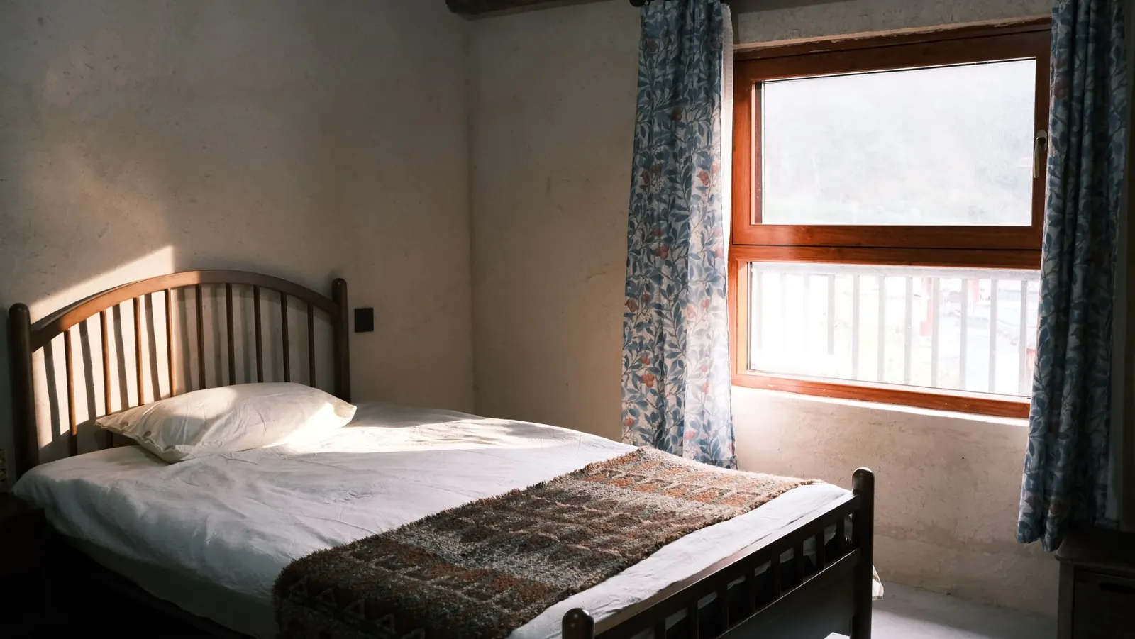 A wooden bed with white sheets against a plain wall, sunlight coming in through a side window with patterned curtains