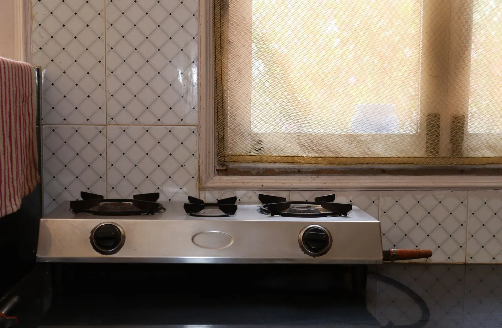 A three-burner steel gas stove on a kitchen counter against a tiled wall, below a window