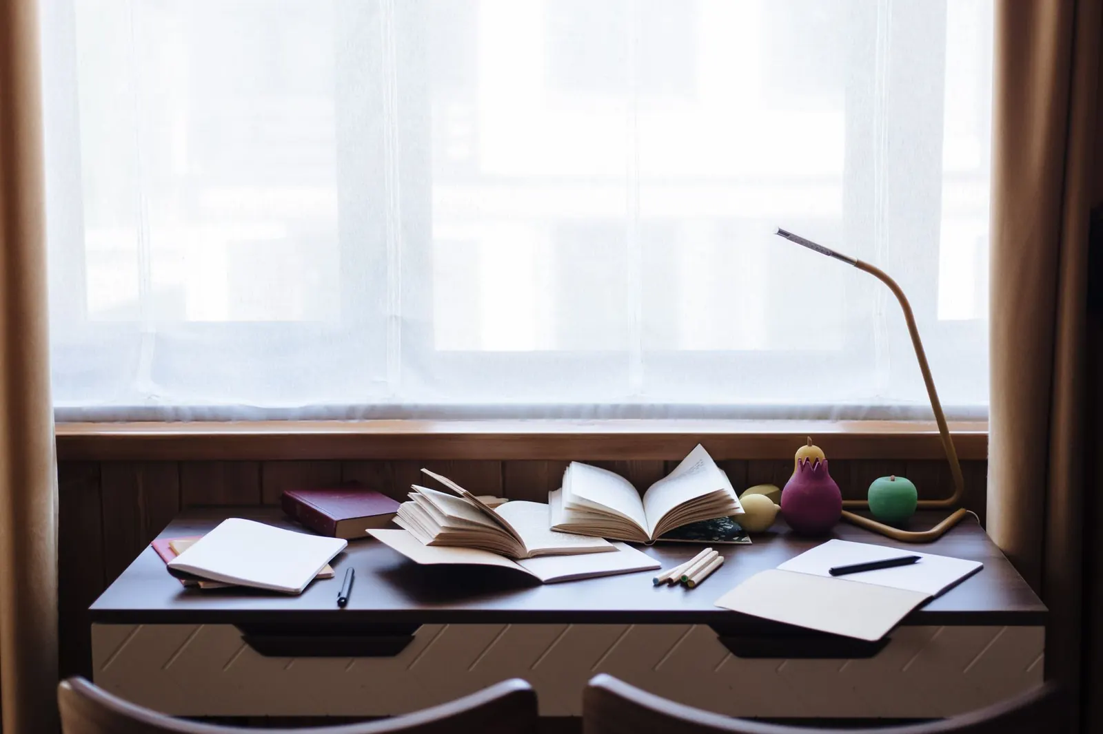 Open books, notebooks and a desk lamp on a study desk set under a curtained window
